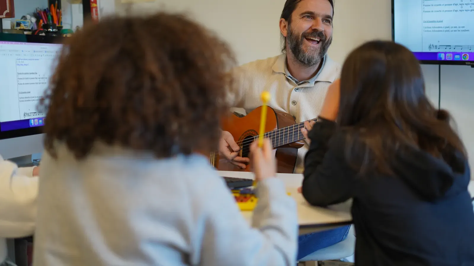 A teacher plays the guitar surrounded by students sitting around a table.