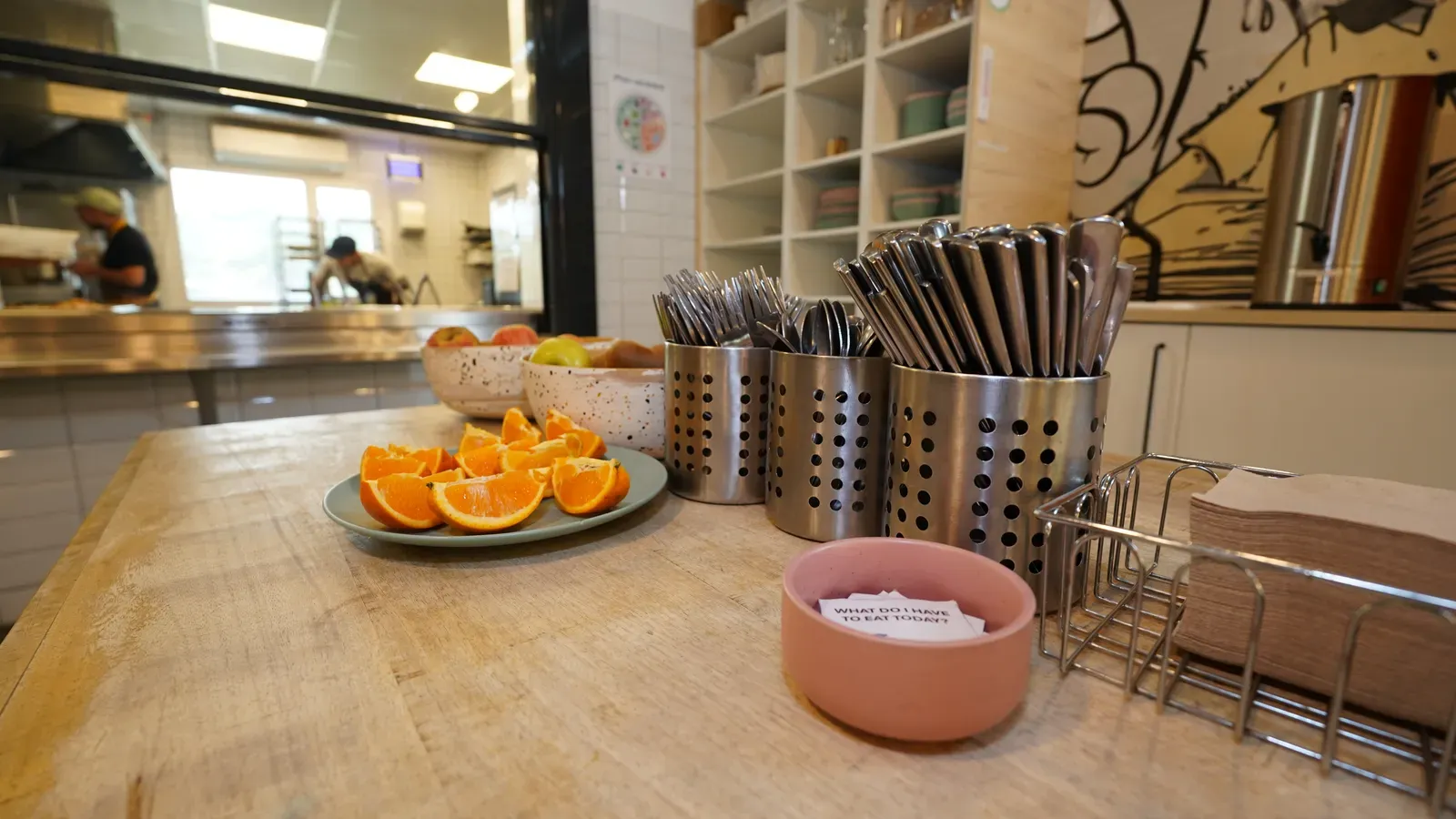 Fruta fresca cortada y cubiertos preparados en el comedor del colegio, con la cocina al fondo.