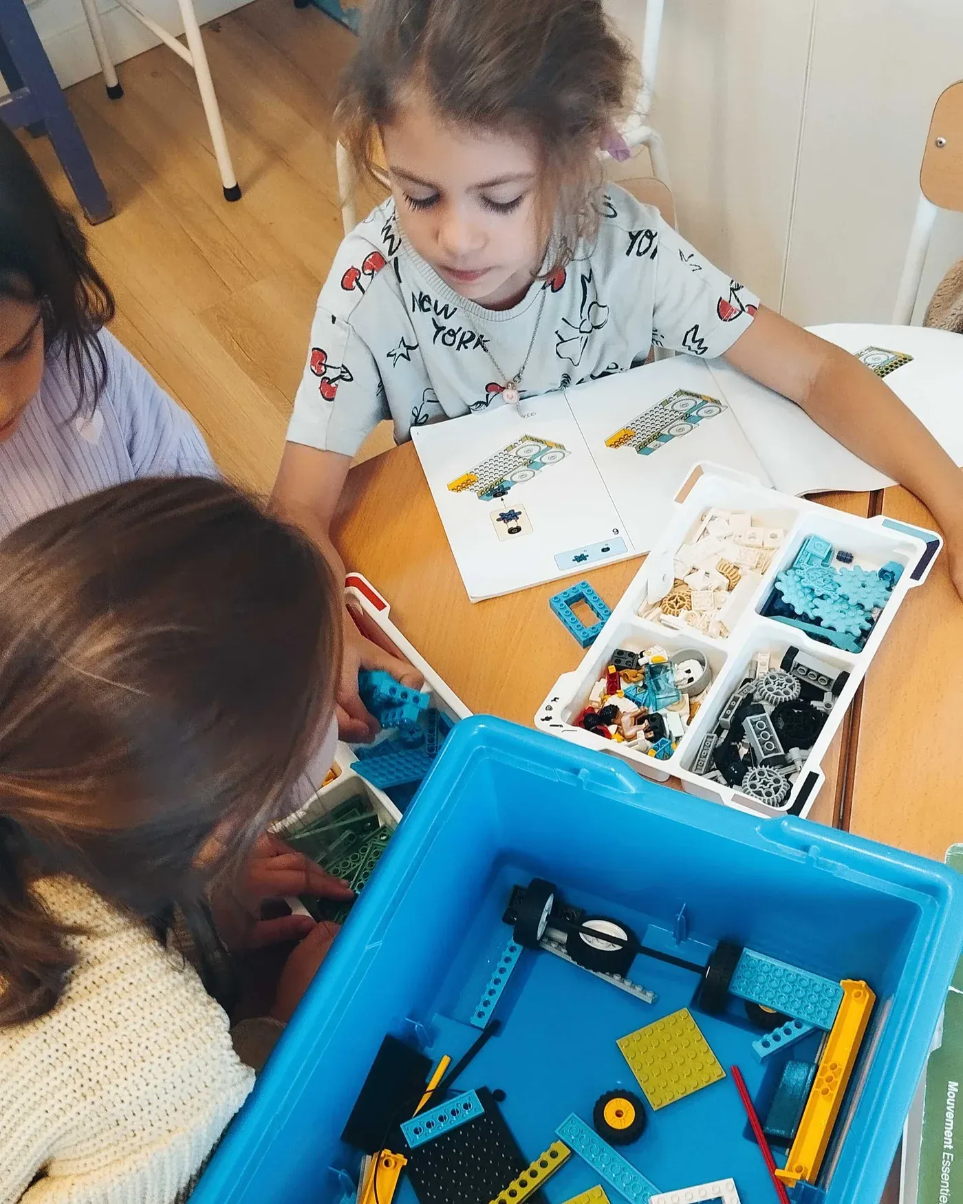 At a classroom table, a pupil follows the building instructions next to her classmates and a box of LEGO bricks