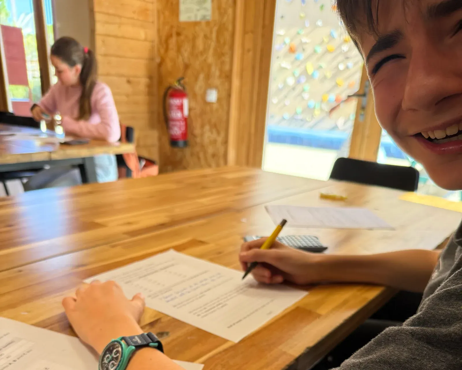Un alumno sonríe a cámara mientras resuelve ejercicios con lápiz y calculadora en una mesa de madera.