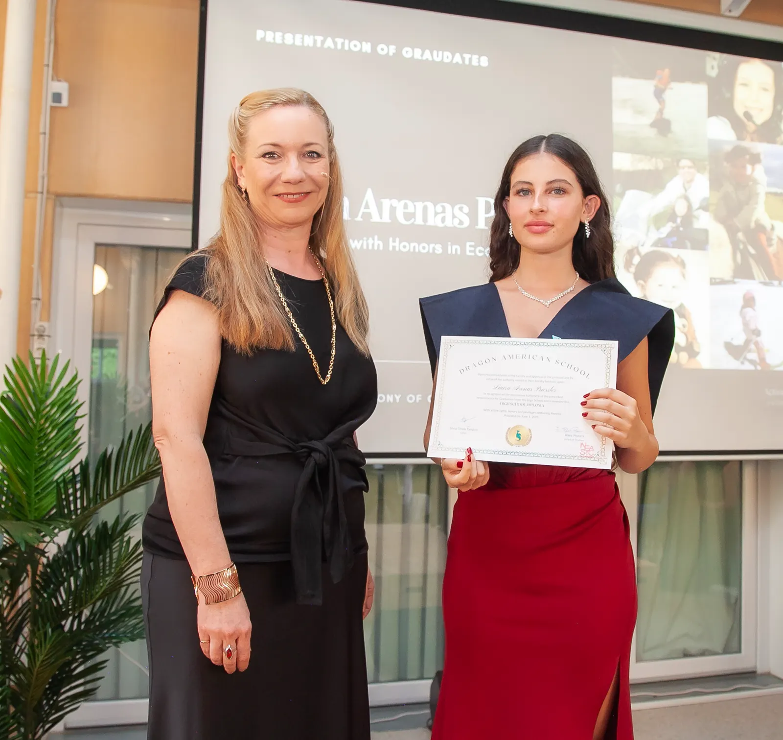 Una alumna recibe su diploma de graduación junto a una profesora, ante la pantalla de la ceremonia.