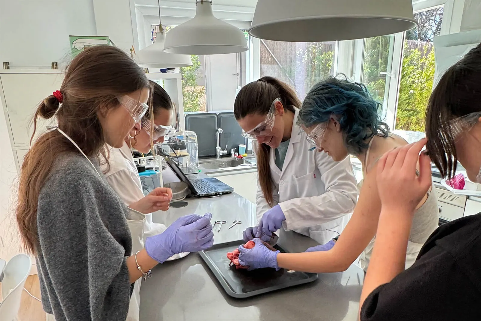 Tres alumnas con bata, gafas y guantes trabajan en una práctica de laboratorio.